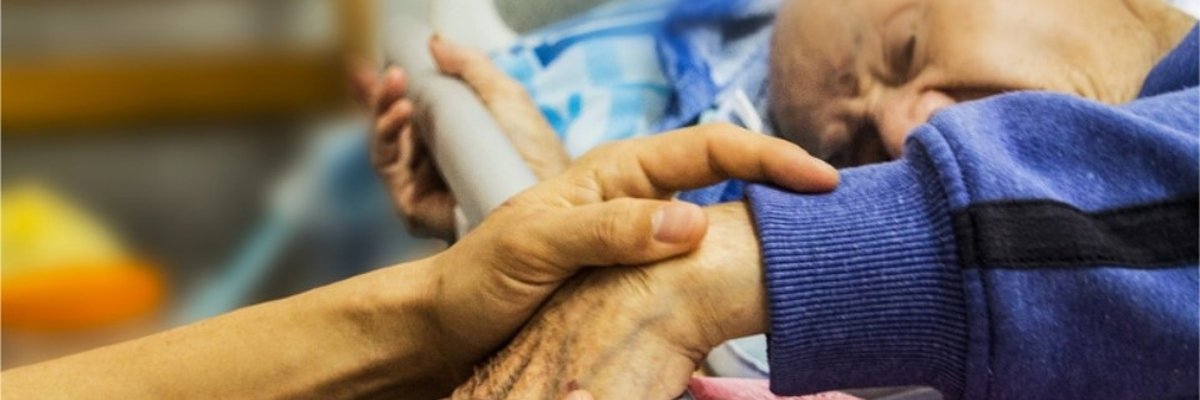 Eine jüngere Person hält einem alten Menschen im Krankenbett die Hand